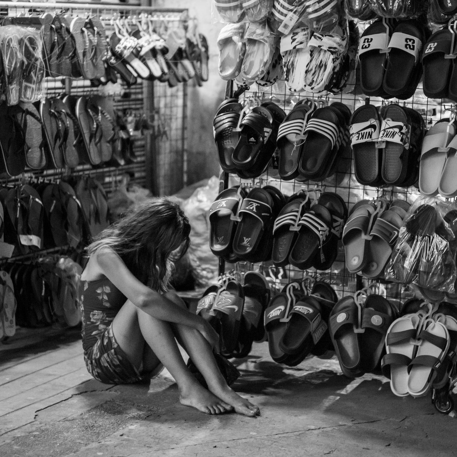 Woman crouches among diverse sandals in a rustic, busy shop.