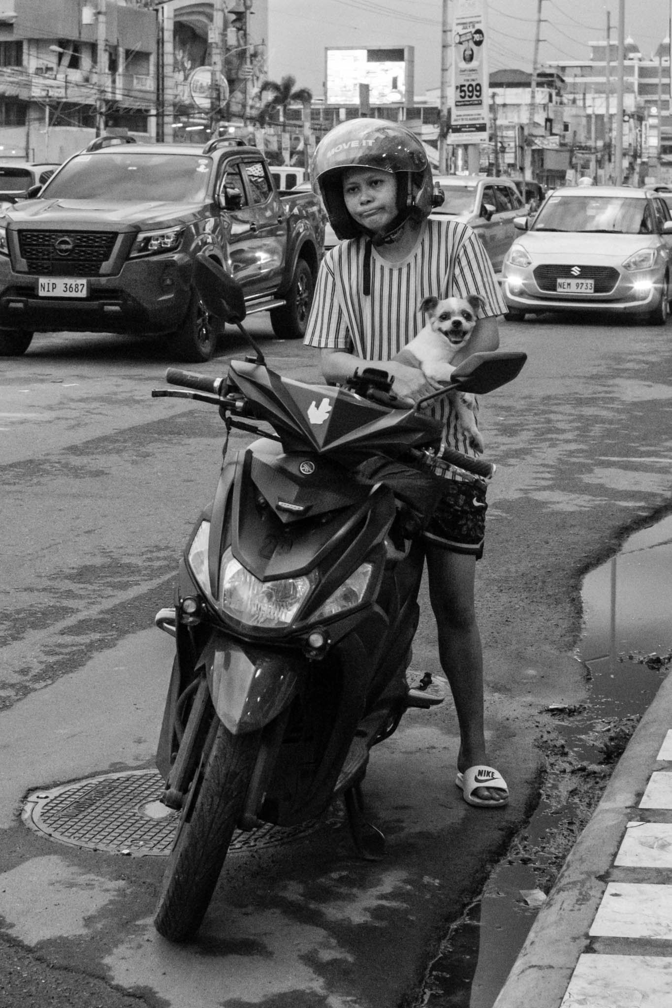 Woman with dog pauses on scooter in busy urban street scene.