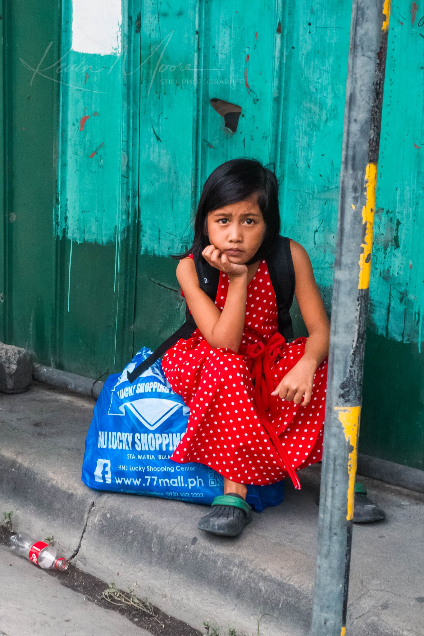 Young girl in the Philippines with a red polka dot dress waits for public transportation with a green backdrop.