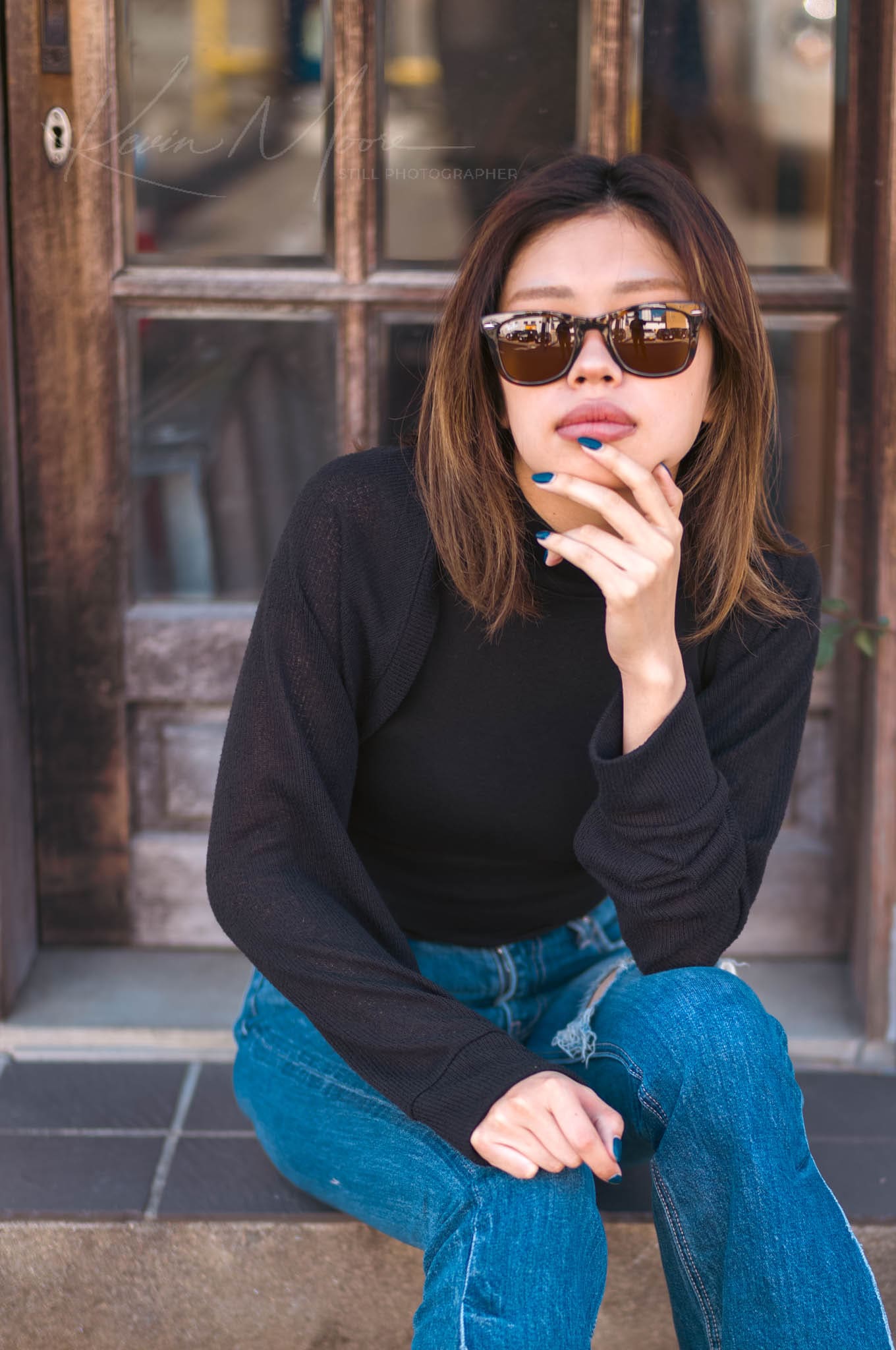 Young woman in sunglasses seated by vintage wooden door, wearing casual black top and blue jeans.