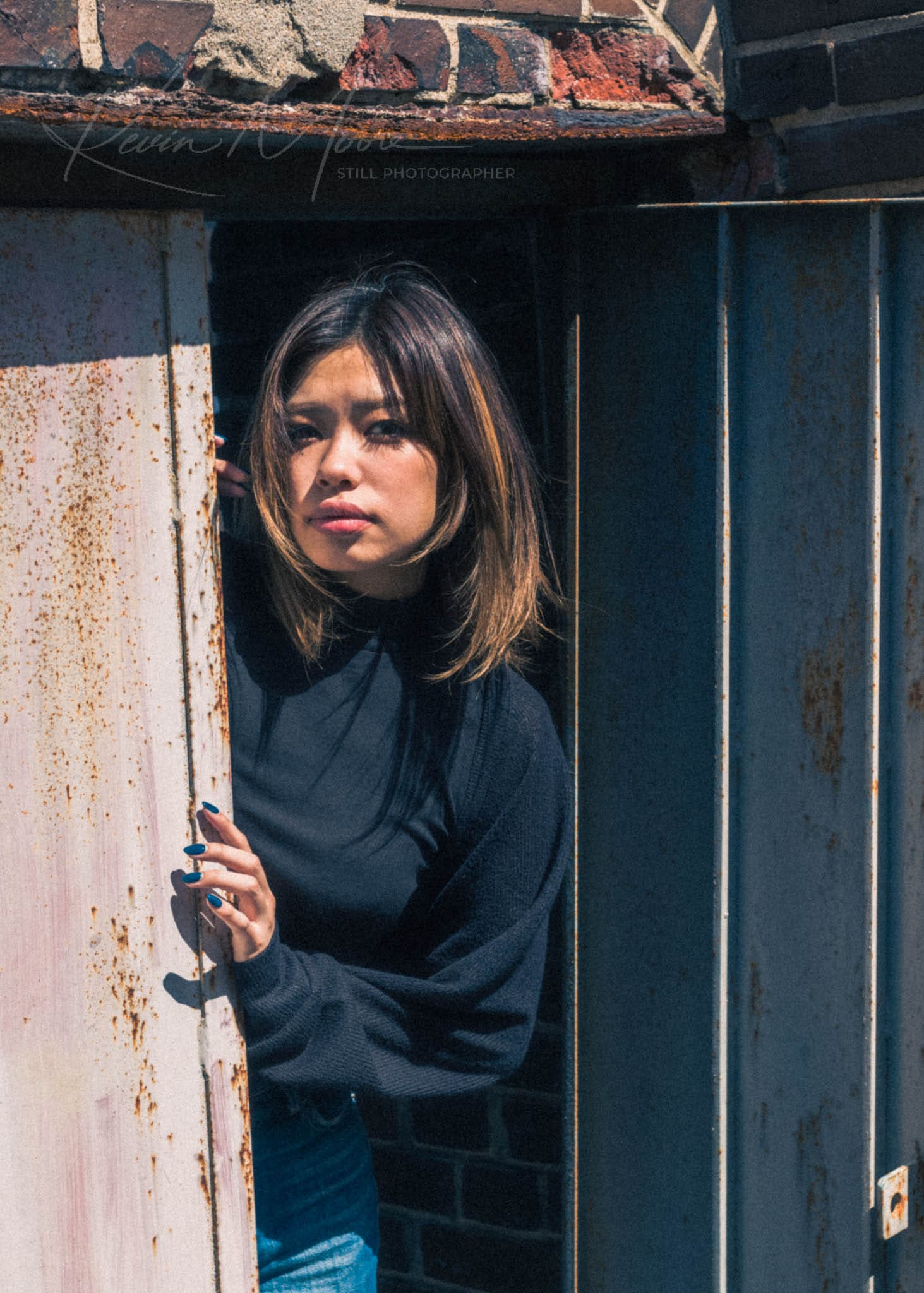 Woman with balayage hair peeking from a rusted metal door in an urban setting.