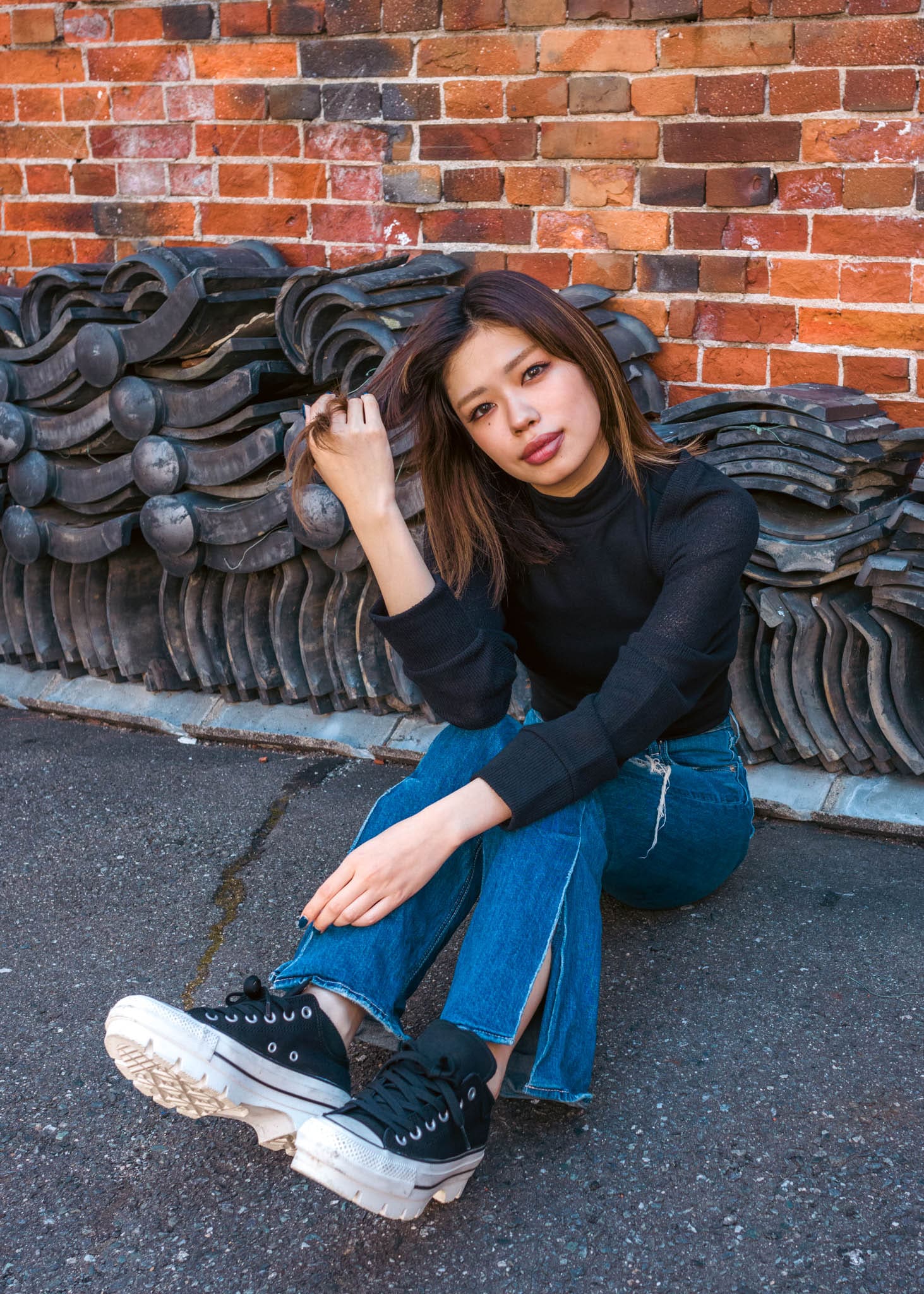 Young woman in black turtleneck and jeans sitting by brick wall with roofing tiles.