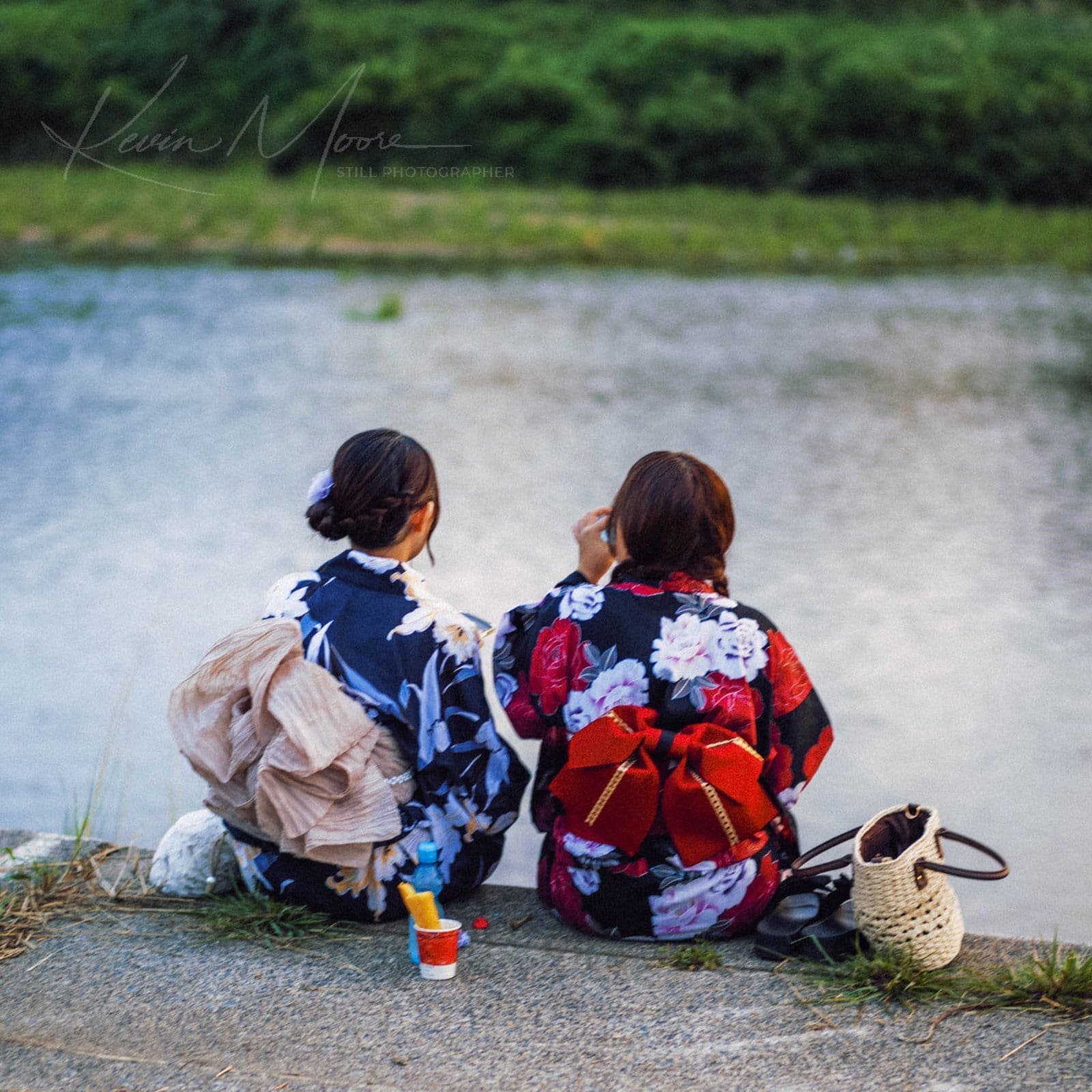 Two women in traditional kimonos sitting by a tranquil river, reflecting cultural heritage.