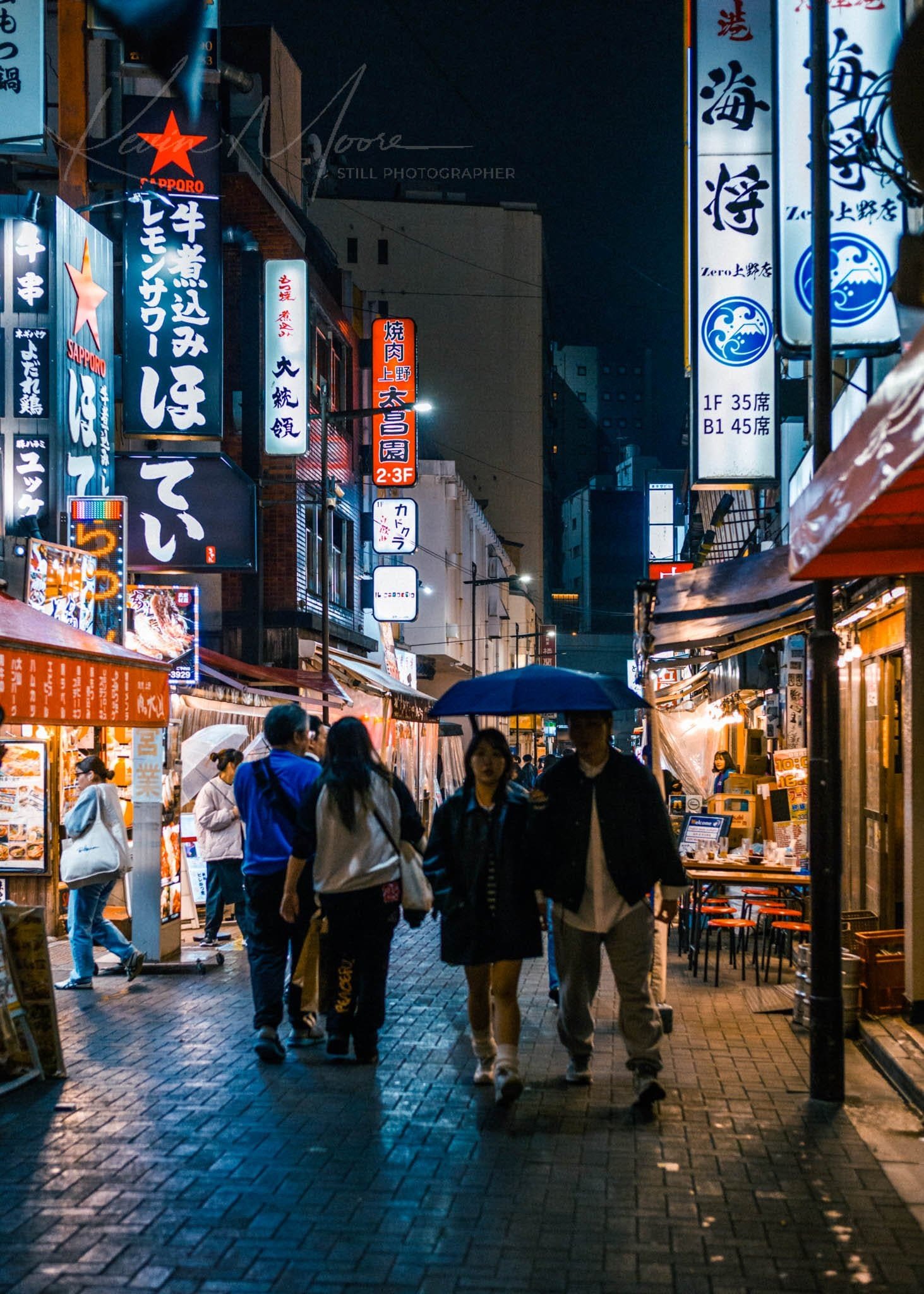 Busy Tokyo street at night with neon signs and people under a blue umbrella.